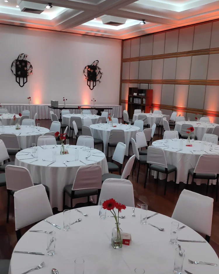 Salão de eventos montado com mesas redondas, toalhas brancas e flores vermelhas